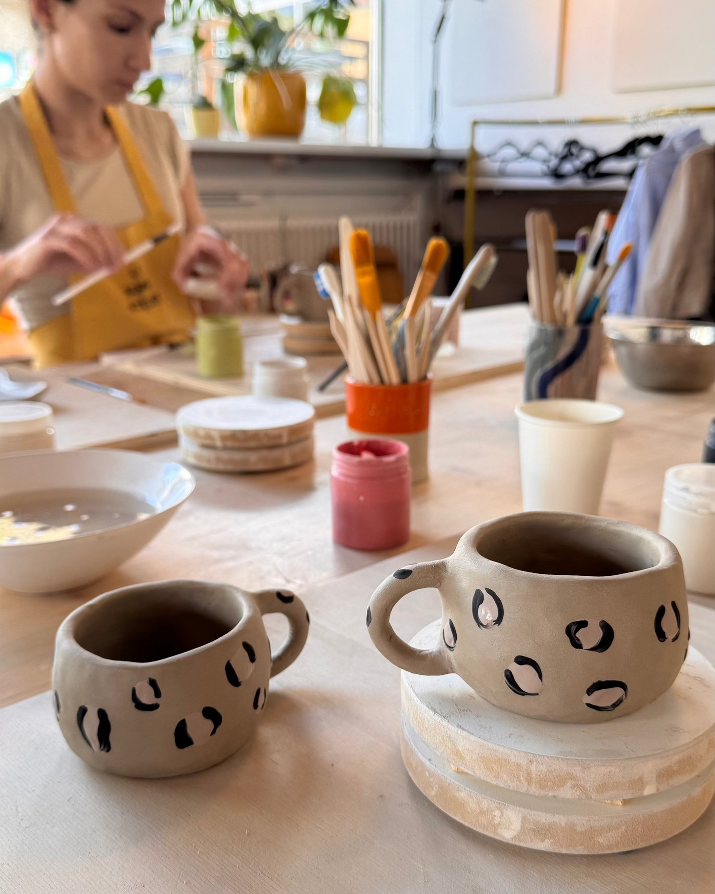 Deux tasses en céramique à motifs léopard sur une table d’atelier, avec une participante peignant en arrière-plan.