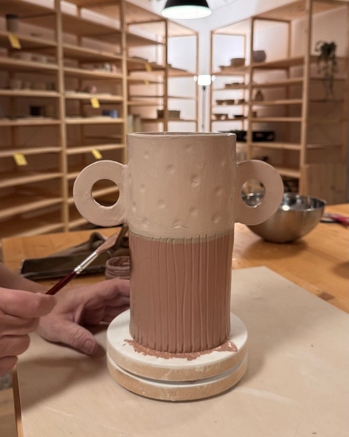 Décoration au pinceau d’un vase en céramique beige et rose à deux anses rondes dans l’atelier.